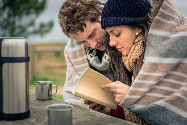 Young couple under blanket reading book outdoors in a cold day