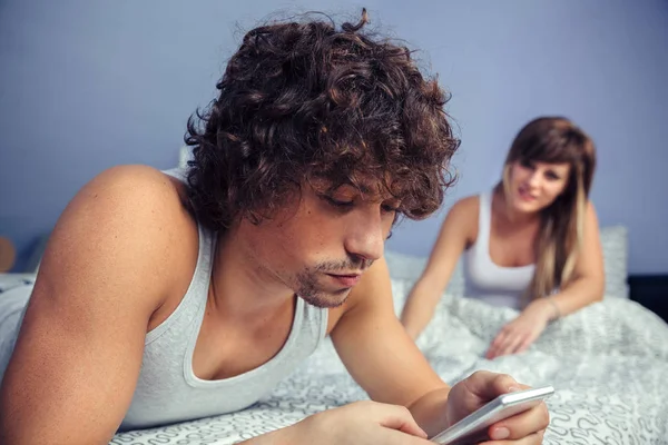 Homem deitado usando smartphone e mulher sentados na cama — Fotografia de Stock