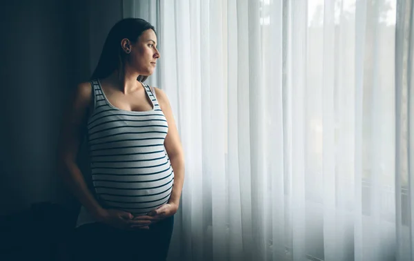 怀孕妇女抱着她的腹部在窗帘前 — 图库照片