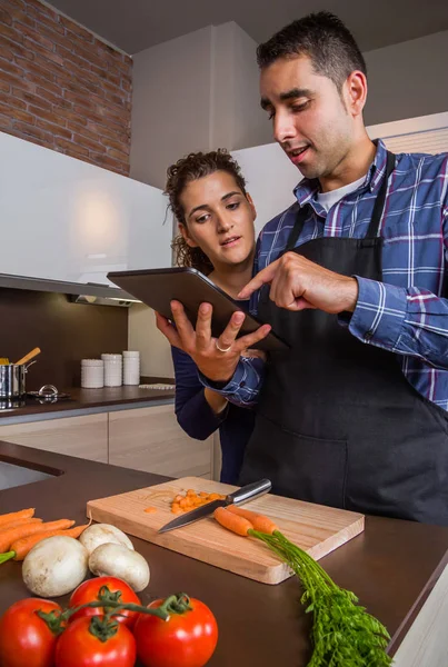 Casal em casa cozinha procurando receita com um tablet eletrônico — Fotografia de Stock