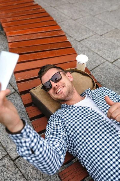 En mann som ligger på en benk og lager selfie med mobilen. – stockfoto