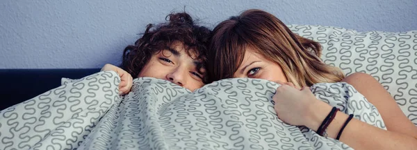 Happy couple laughing and covering mouths under duvet — Stock Photo, Image