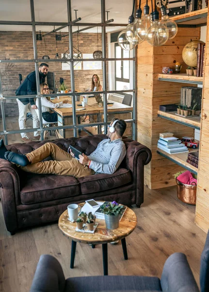 Businessman lying on an office sofa talking to his coworkers — Stock Photo, Image