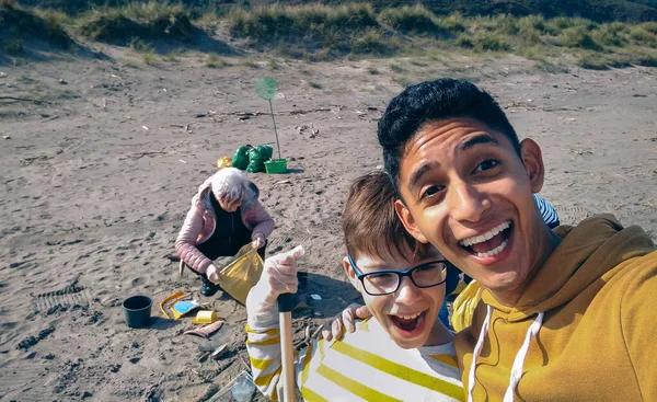 Молодой человек и мальчик делают селфи, пока группа добровольцев убирает пляж — стоковое фото