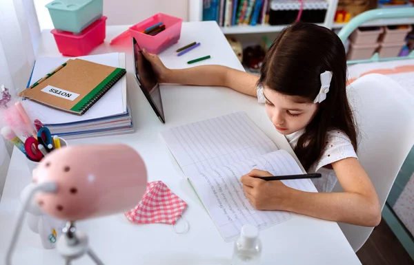 Chica estudiando en casa con la tableta y la máscara en la mesa —  Fotos de Stock