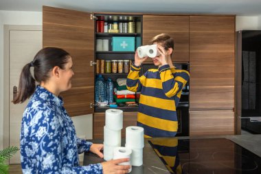 Neşeli evlat tuvalet kağıtlarını dürbün olarak kullanırken gülümseyen anne de kilerde acil durum malzemelerini düzenliyor. Potansiyel felaketler için aile hazırlığı. 72 saat hayatta kalma kiti.