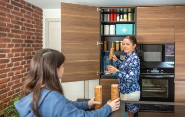 Anne ve kız, 72 saatlik acil durum çantasına kurabiye paketleri ekliyor. Savaş ya da yiyecek sıkıntısına karşı hazırlar. Aile, mutfakta acil durum kileri hazırlamak için işbirliği yapıyor.