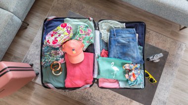 Sehpanın üzerine renkli giysiler ve aksesuarlarla yerleştirilmiş açık bavul manzarası, tatile hazırlanmak, klasik fotoğraf makinesi ve sarı güneş gözlüğünün yanında