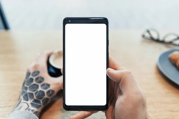 Primer plano del hombre tatuado manos sosteniendo el teléfono móvil con pantalla en blanco . — Foto de Stock