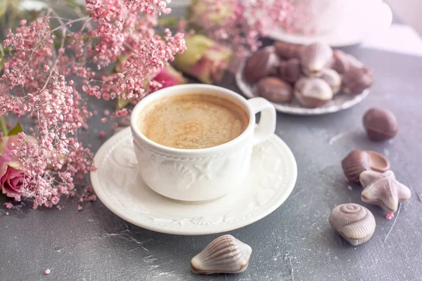 Eine Tasse duftenden Kaffee am Morgen und einen Strauß rosa Blumen. Schokoladenbonbons in Form von Herzmuscheln. Muscheln auf den Tassen. Romantik und helle Farben. — Stockfoto