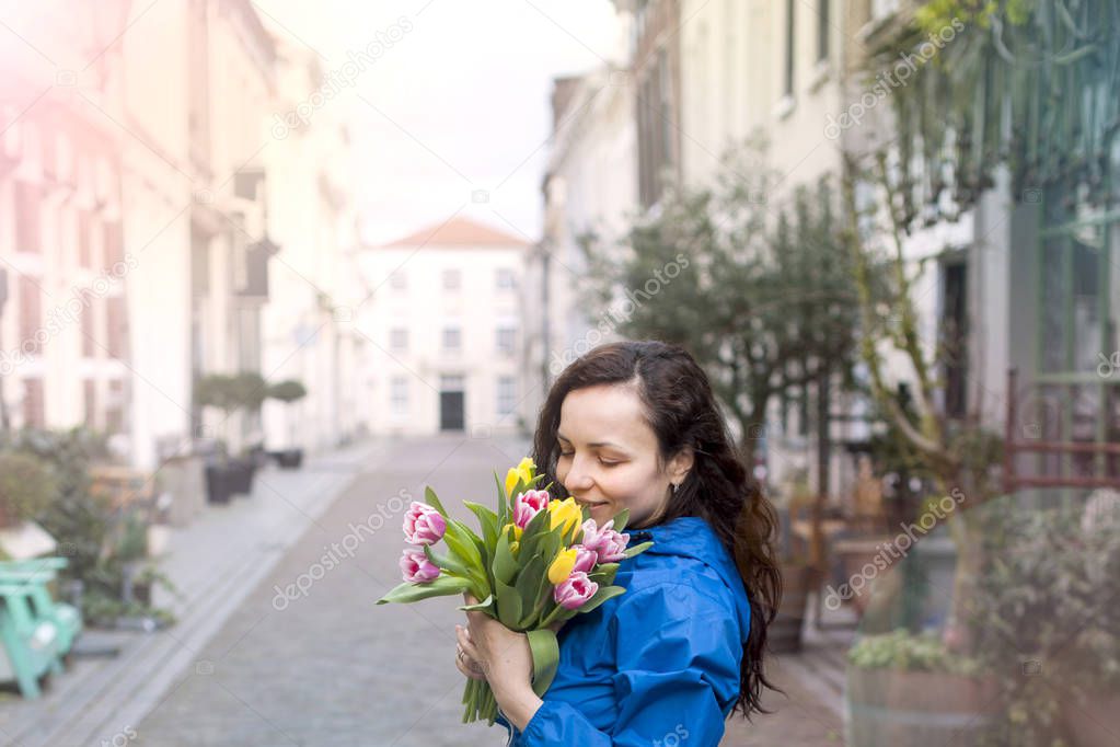 Happy Woman with a bouquet of fresh tulips yellow and pink in the hands. With a basket for shopping. On the street of the old European city. Blooming flowers. Free space for text