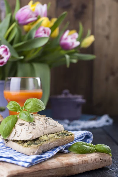 Panini con basilico e formaggio. Pane con cereali. Un bicchiere di succo di carota. Colazione sana. Tovagliolo in una gabbia blu e posto per il testo. Bouquet di tulipani sul retro  . — Foto Stock