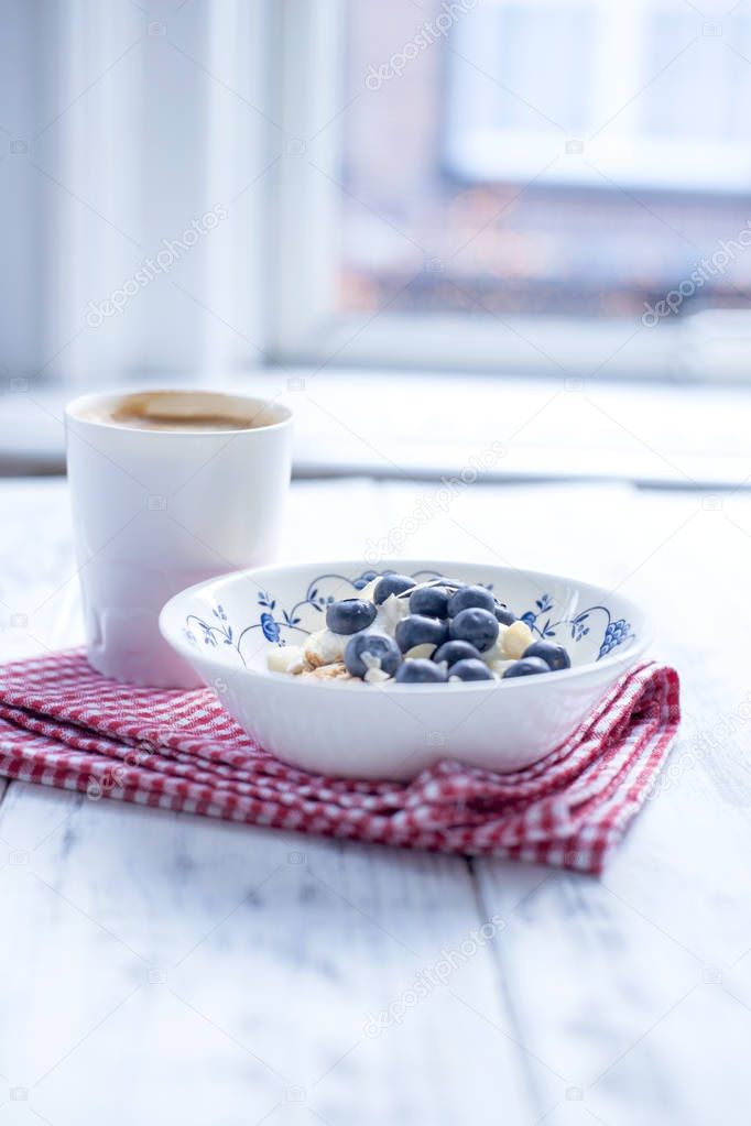 Plate with muesli and berries, coffee in a cup for breakfast. Napkin in a red cage on a white wooden table by the window. Free space for text