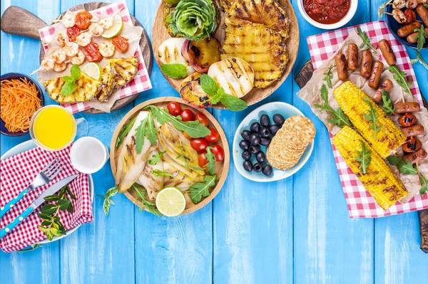 Grilled vegetables, shrimp, fruit on a wooden plate and sausages, juice and salad on a blue background. Summer dinner. Free space for text. Copy space