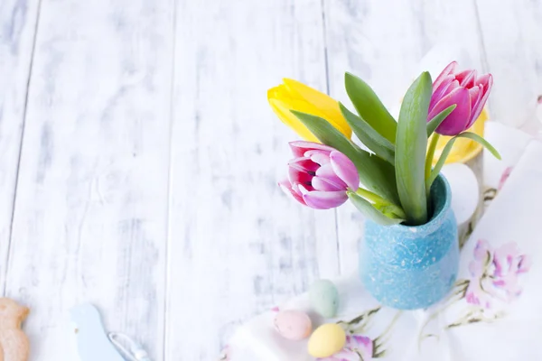 Spring tulips are pink and yellow. Different colors of eggs and pottery rabbit. Decor for Easter. White wooden background and place for text
