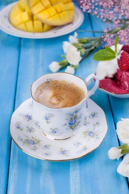 Hoş kokulu kahve bir güzel porselen fincan mavi çiçekler ve meyveli dondurma ile. Çiçekler ve olgun mango mavi ahşap arka plan üzerinde. Kahvaltı tatlıdır