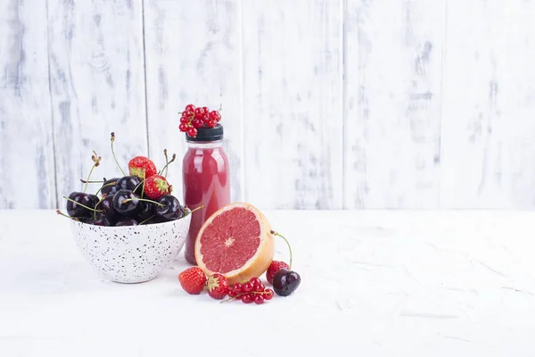 Una taza con cerezas maduras, pomelo, frambuesas, una botella de jugo y un fondo blanco. Bayas de verano. Copiar espacio —  Fotos de Stock