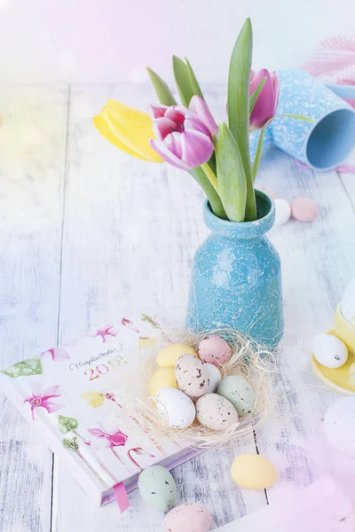 The tulips are pink and yellow in a vase on the table. White background. Free space for text or a postcard. Easter. Notepad with flowers. Easter bunny and eggs are colored.