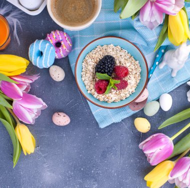 Gri taş zemin üzerine lezzetli bahar kahvaltı. Bir buket renk pembe ve nane taze lale. Küçük ve büyük renkli Paskalya yumurtaları. Yulaf ezmesi ahududu meyveleri, bisküvi, kahve. Kartpostal