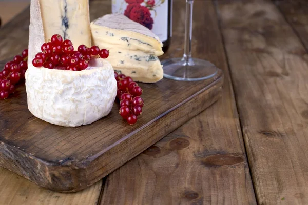 A bottle of red wine and a glass. Assorted soft cheese with mildew. Blue cheese. Fresh berries of red currant. Branch of spring white flowers. Wooden brown background. Free space for text or advertisi