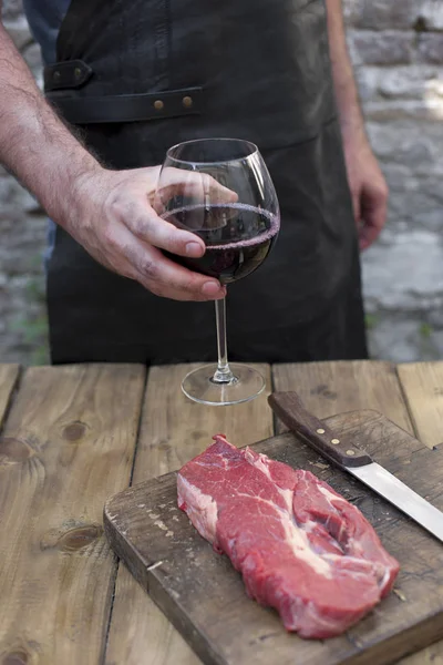 Una copa de vino tinto en las manos de un hombre en un delantal de cuero negro. Un hombre está cocinando carne. Copiar espacio —  Fotos de Stock