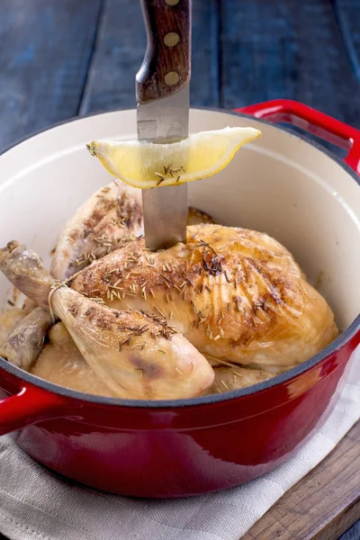 The whole chicken baked with lemon and rosemary in a red cast iron. Blue wooden background and gray towel. The knife is stuck in the chicken and fork. Free space for text