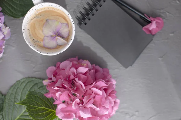 Uma xícara de café perfumado da manhã e flores de hortênsia rosa, em um fundo cinza, espaço livre para texto e um bloco de notas preto. Cartão, espaço de cópia — Fotografia de Stock