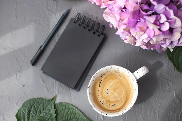 Uma xícara de café perfumado da manhã e flores de hortênsia rosa, em um fundo cinza, espaço livre para texto e um bloco de notas preto. Cartão, espaço de cópia — Fotografia de Stock