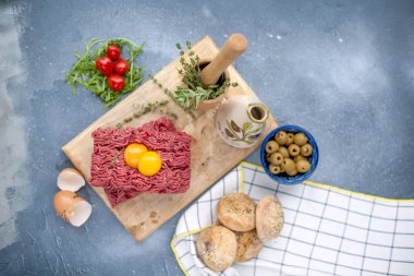 Taze et et forcemeat, ahşap oyun tahtası olarak. Çiğ yumurta ve domates küçük vardır. Otlar pişirme için ve yuvarlak çörekler. Bir gri taş arka plan üzerinde. Metin yazmak için boş alan.