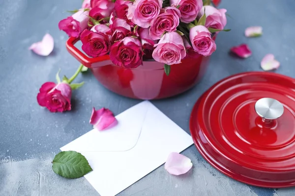 Un bouquet de roses fraîches, dans une casserole de fonte rouge, sur un fond de pierre grise. Près de la fenêtre. Une enveloppe blanche. Endroit gratuit pour écrire du texte ou une carte postale — Photo
