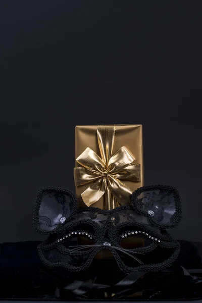 Gift in gold paper with a bow and carnival mask on a black background — Stock Photo, Image