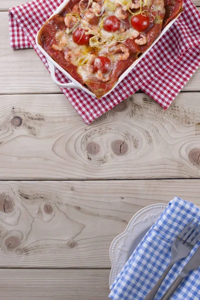 Lasagna baked in a white baking sheet, on a napkin in a red cage and a plate with a blue napkin on a wooden background. — Stock Photo, Image