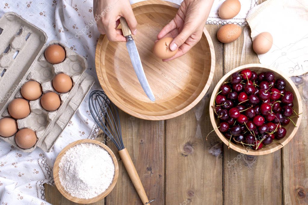 Female Hands break the egg into a wooden bowl. Preparation of dough for a pie with a cherry. Summer homemade pastries. Top view. Copy space