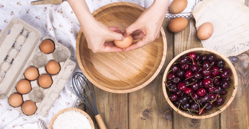 Female Hands break the egg into a wooden bowl. Preparation of dough for a pie with a cherry. Summer homemade pastries. Top view. Copy space