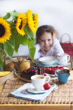 Küçük kız evde kahvaltı. Bir buket çiçek ayçiçeği ve tatlı bir pasta meyve, ev yapımı hamur işleri ve çilek ve kahve ile geçerli. Mutlu bir çocuk. Kopya alanı