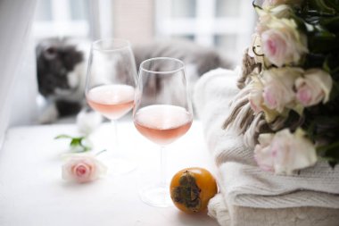 İki bardak şarap ve kış sıcak giysiler evde bir sürü pencere yakın. Kedi, büyük buket gül ve çerçeve içinde sonbahar meyve. Pencerenin dışında şehirdir. Metin için boş alan.