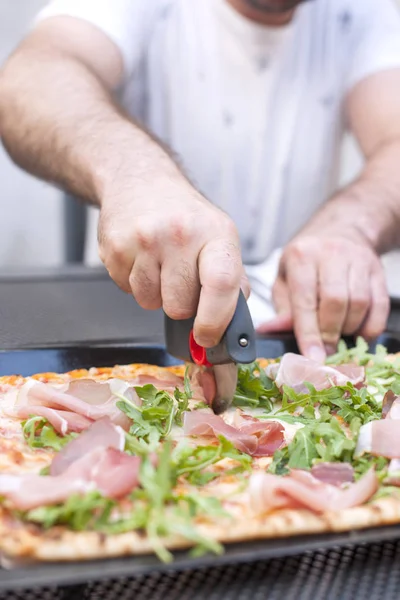 Talian pizza met rucola en ham. Heerlijke traditionele gerechten voor de lunch. De hand snijdt de pizza in gedeelten. Homemade bakken. Bovenaanzicht. — Stockfoto