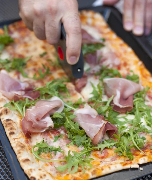 Talian pizza with arugula and ham. Delicious traditional food for lunch. The hand cuts the pizza into portions. Homemade baking. Top view. — Stock Photo, Image