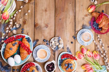 Paskalya kahvaltısı taze kahve, böğürtlen ve hamur işleri, turuncu laleler, pastırmalı kruvasanlar ve çeşitli tatlılar, renkli bıldırcın yumurtaları ve bahar tatili süslemeleriyle yatıyordu. Üst Manzara. Boşluğu kopyala