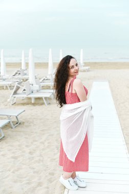 Kırmızı elbiseli ve kıvırcık saçlı bir kadın sahilde duruyor.. 