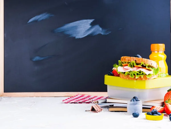 Caja con almuerzo escolar cerca de la pizarra negra. Comida saludable para un niño. Tostadas con ensalada y fresas y arándanos y una botella de jugo. Comida y libros para el instituto o escuela. Copiar espacio —  Fotos de Stock