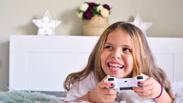 Una niña está jugando una consola de ordenador en un sofá en una habitación luminosa. Juegos simultáneos para niños. Joystick para control en las manos. — Vídeos de Stock