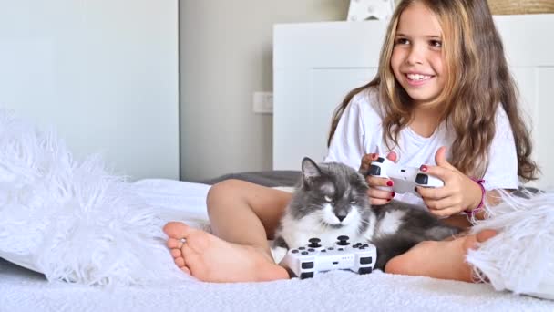 Una niña está jugando una consola de ordenador en un sofá en una habitación luminosa. Juegos simultáneos para niños. Joystick para control en las manos. Niño alegre y un gato. Copiar espacio. Imágenes de alta calidad 4k. — Vídeos de Stock