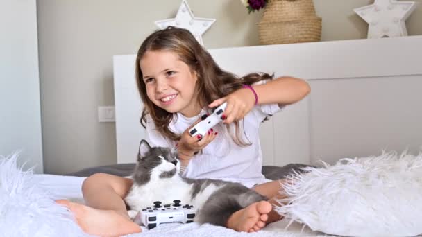 Une petite fille joue une console d'ordinateur sur un canapé dans une pièce lumineuse. Jeux simultanés pour les enfants. Joystick pour le contrôle dans les mains. Enfant joyeux et un chat. Espace de copie. Images 4k de haute qualité — Video