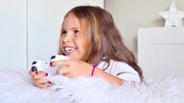Küçük bir kız parlak bir odada koltukta bilgisayar konsoluyla oynuyor. Çocuklar için eş zamanlı oyunlar. Kontrolü ele almak için joystick. Çocuğun duyguları ve eğlencesi. Uzayı kopyala Yüksek kalite 4k görüntü