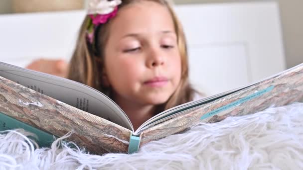 Die Schülerin liest. Kleines Mädchen beim Lesen eines Buches im Schlafzimmer auf dem Bett. Bildung und Hobbys der Kinder. Tageszeit und helle Farben. — Stockvideo