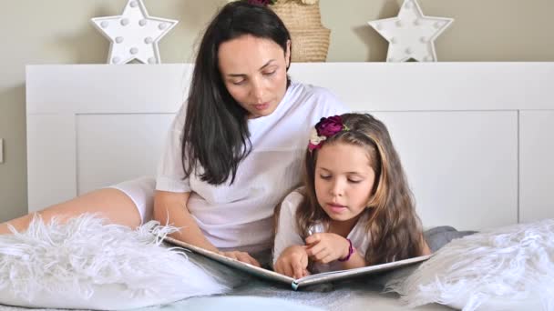 Mamá lee un libro con su hija. Niña, la colegiala está leyendo en el dormitorio de la cama. Comunicación entre hijos y padres. Colores diurnos y claros en el marco. Enfoque suave. — Vídeos de Stock