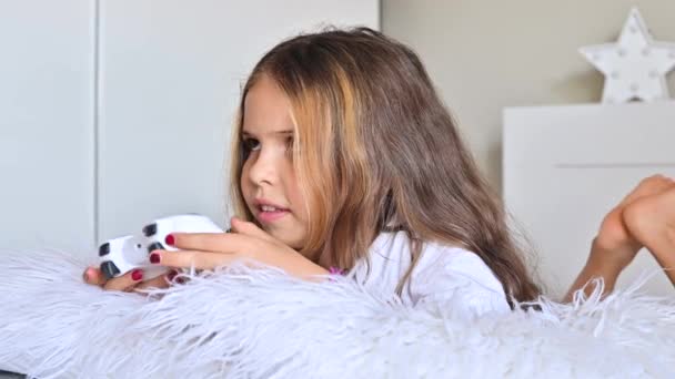 Uma menina está jogando um console de computador em um sofá em uma sala brilhante. Jogos simultâneos para crianças. Joystick de controle em mãos. Emoções e diversão da criança. Espaço para cópia. Imagens 4k de alta qualidade — Vídeo de Stock