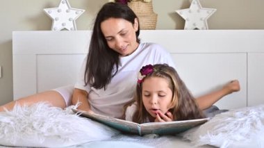 Annem kızıyla birlikte bir kitap okuyor. Küçük kız, okul kızı yatak odasında yatağın üstünde kitap okuyor. Çocuklar ve ebeveynler arasındaki iletişim. Çerçevede gündüz ve açık renk var. Yumuşak odak.