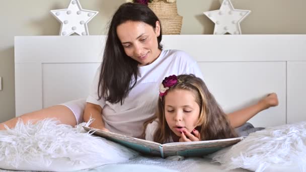 Mamá lee un libro con su hija. Niña, la colegiala está leyendo en el dormitorio de la cama. Comunicación entre hijos y padres. Colores diurnos y claros en el marco. Enfoque suave. — Vídeos de Stock